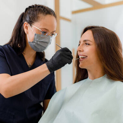 A dental hygienist wearing gloves and a mask is checking a patient's teeth using a dental instrument. The patient is seated comfortably in a modern clinic, smiling during the examination.